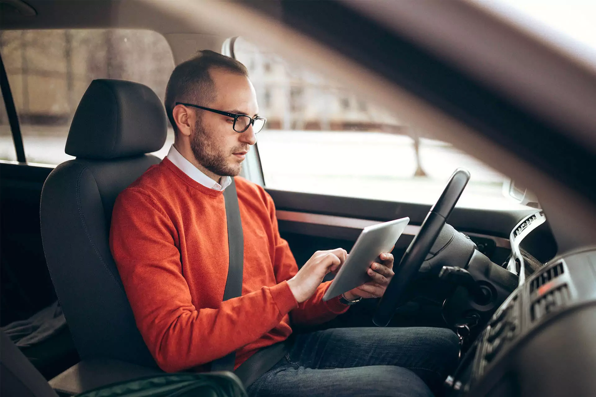 Man in orange sweater scrolling on a tablet in driver’s seat of a parked car