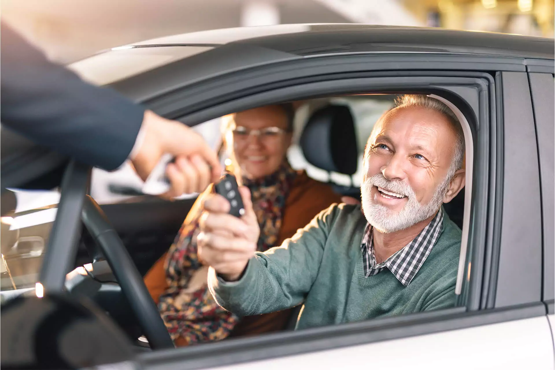 Smiling old man in driver’s seat of a car receiving car keys with woman in passenger seat