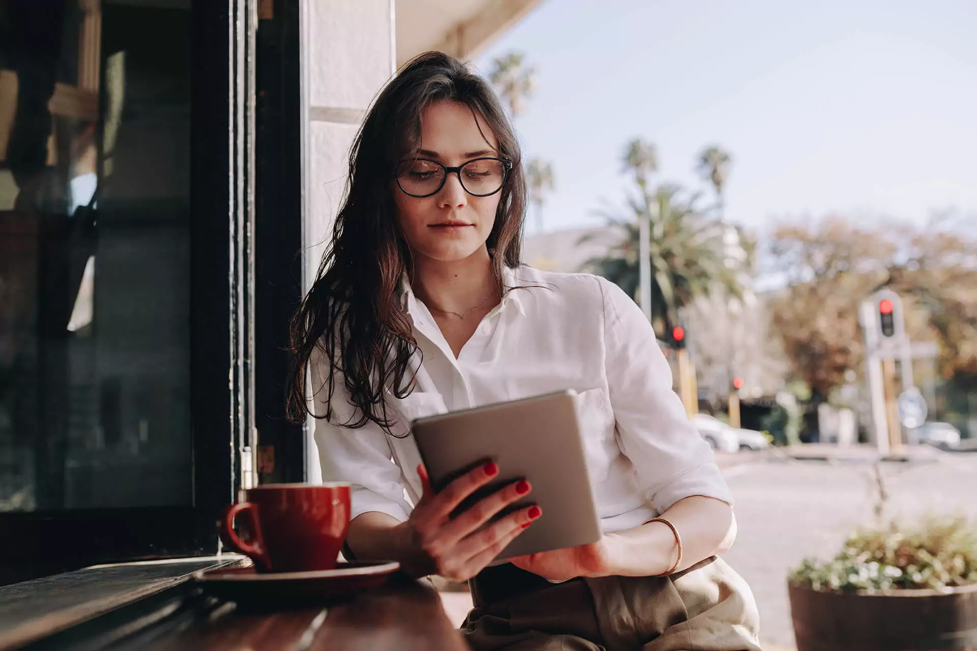 Woman holding tablet sitting at an outdoor cafe table with coffee cup on it