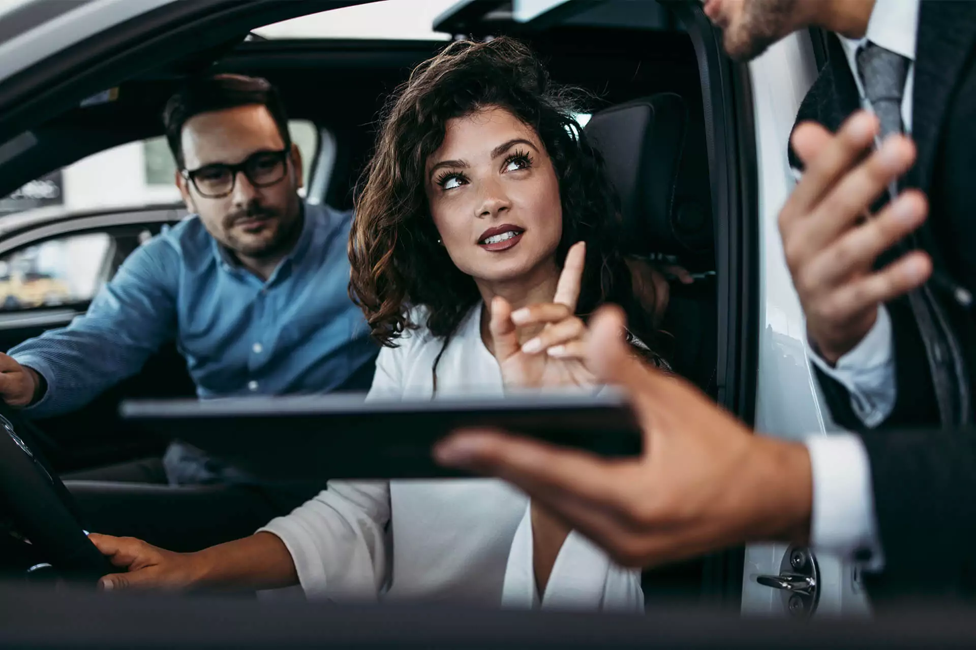 Woman in driver’s seat of a car looking at a salesman’s tablet with man in passenger seat