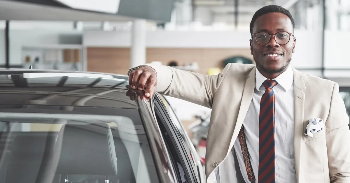A dark-skinned man in a beige suit with a striped tie and suspenders, standing beside a black car in a bright, modern car showroom. He is smiling confidently, leaning on the car's roof and driver's side door frame, with a professional and approachable demeanor.