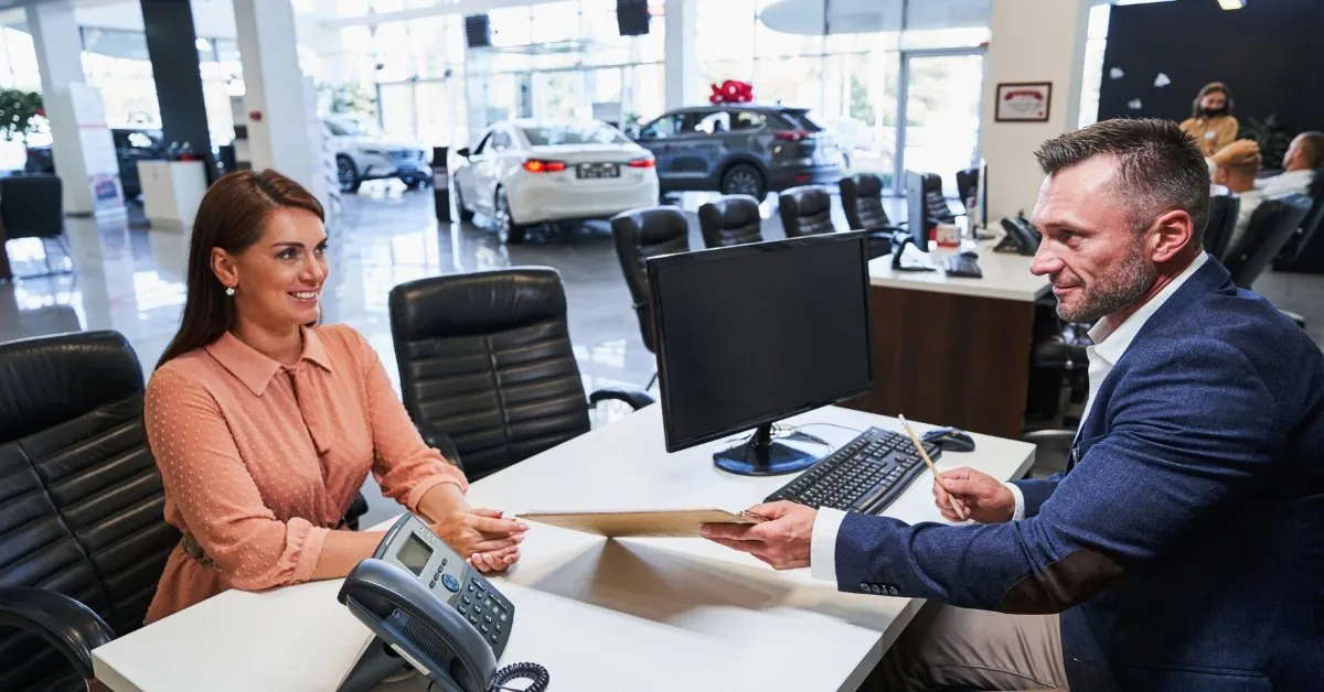 A car dealership scene showing a smiling female customer seated at a desk, receiving documents from a male salesperson or finance manager. The desk features a computer monitor, keyboard, and phone. In the background, a showroom displays new cars, including a dark grey SUV with a red bow and a white sedan, under bright natural lighting.