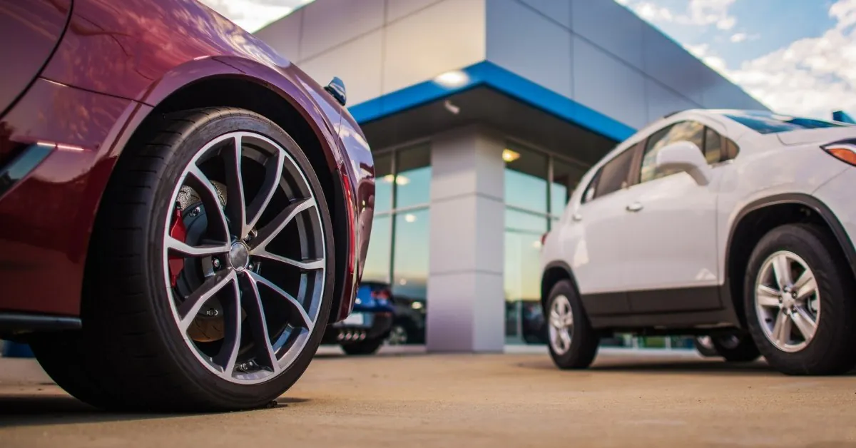 Close-up view of a maroon sports car's rear wheel and fender with a red brake caliper, parked in front of a modern car dealership building. A white SUV is visible in the background under a partly cloudy sky.