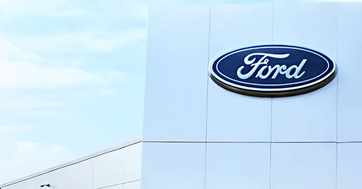 A bright, sunny image of a modern white building with the iconic blue oval Ford logo prominently displayed on its upper right facade. The building features a minimalist design with panel lines forming a grid pattern. The background shows a light blue sky with soft, wispy clouds, enhancing the clean and modern aesthetic.