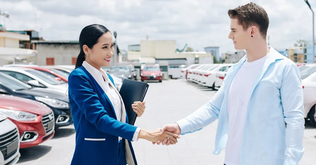 An Asian woman in a dark blue blazer and a Caucasian man in a light blue shirt shake hands in an outdoor car dealership. Rows of cars, including red, black, and white models, are visible in the background under a partly cloudy sky.