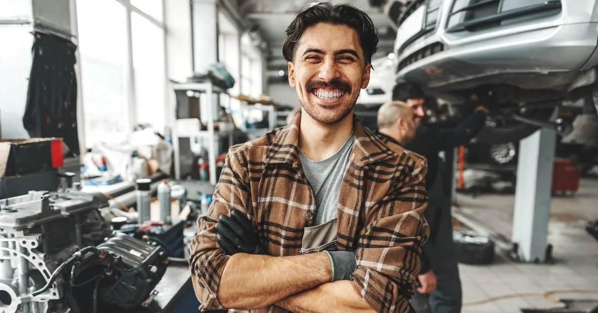 Smiling mechanic with crossed arms in the foreground of a bright auto repair shop, with other technicians servicing a raised vehicle in the background.
