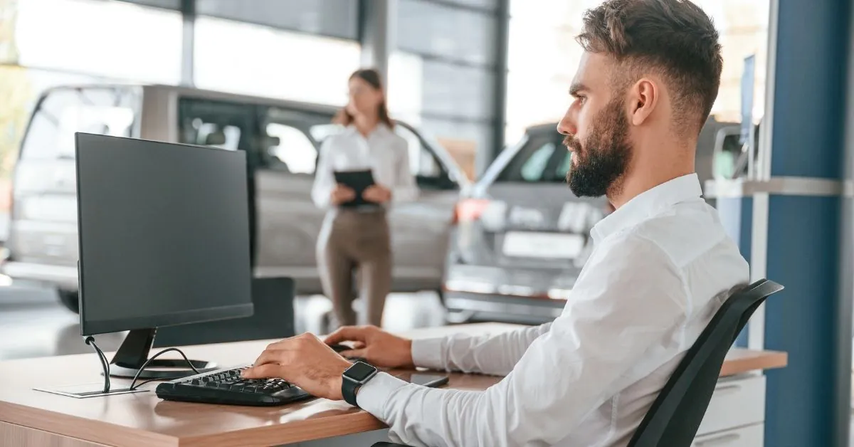 A dealership employee in a white shirt works at a computer desk inside a modern car showroom, with cars and a colleague blurred in the background.